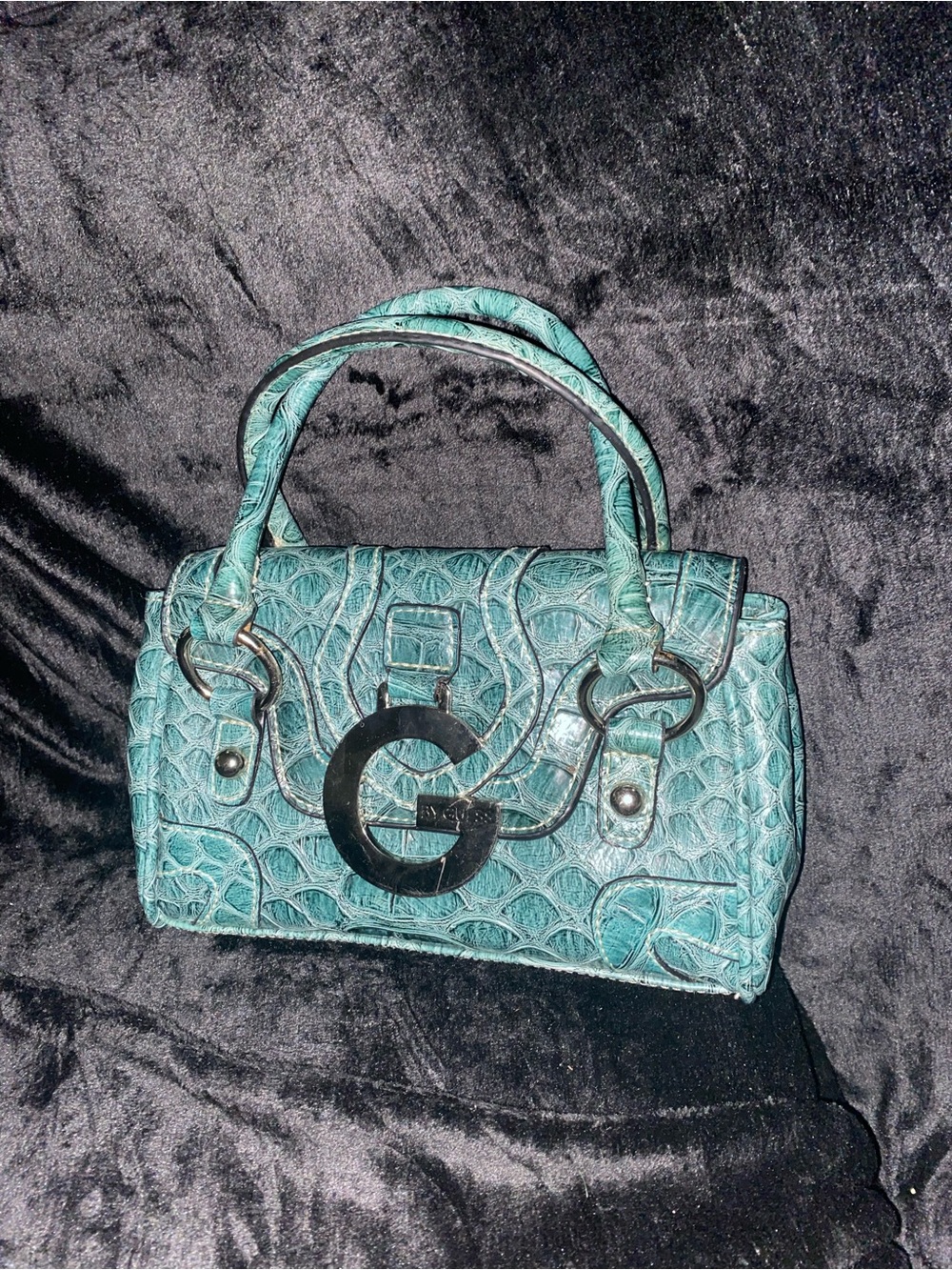 A teal faux-snakeskin textured Guess handbag with 'G' logo hardware.